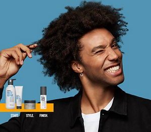 A man with coily hair pulling at a lock of his hair and grinning